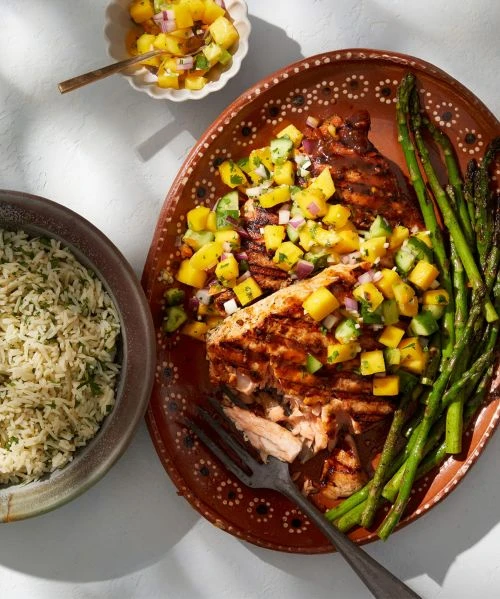 Grilled salmon steak on terracotta plate, topped with diced mango salsa, served alongside cooked asparagus and rice with herbs.
