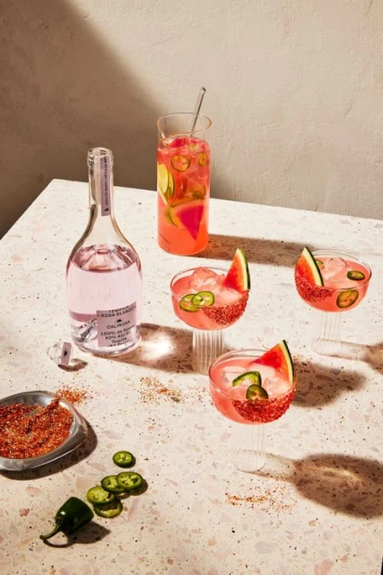 Pink tequila bottle and glasses of mixed beverages with watermelon and jalapenos on a terrazzo surface in a brightly lit indoor setting.