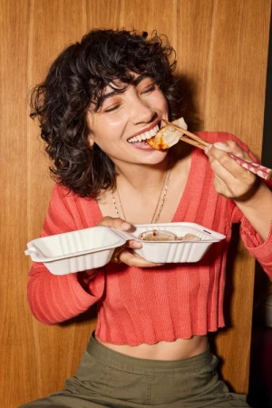 Smiling woman with curly hair, holding takeout container, eating dumplings with chopsticks against a wood-panelled backdrop.