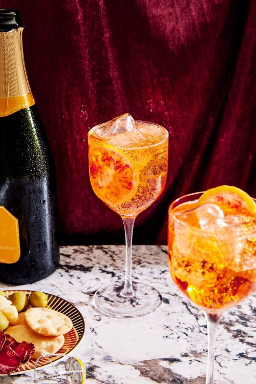 Two glasses of Aperol spritz with ice and orange slices on a marble table next to a bottle of prosecco and snacks.