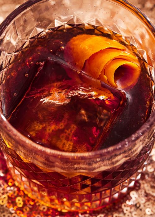 Close-up of a crystal glass containing a dark cocktail with an ice cube and orange peel garnish, set on a shimmering gold surface.