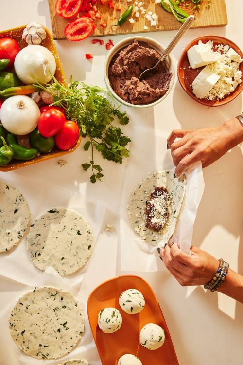 Woman preparing tortillas with beans and cheese at a table with tomatoes, onions, peppers, and tortillas in the background.