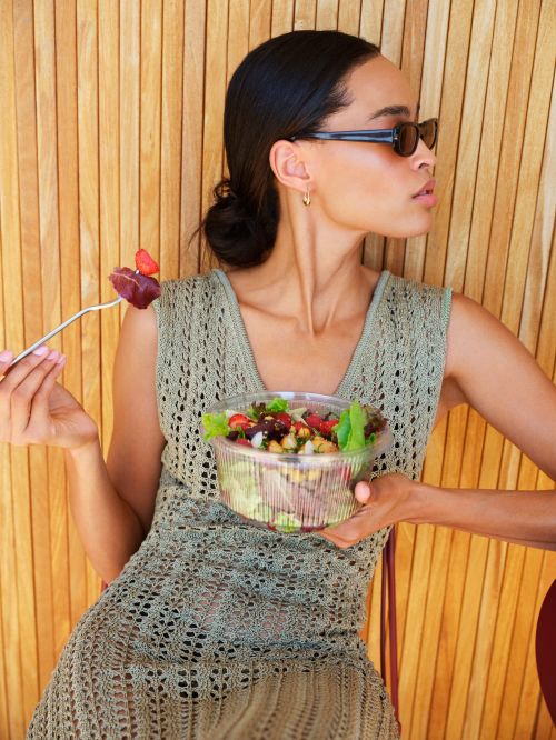 A young woman sits in front of a wooden wall, eating a salad. She's wearing sunglasses and a knit dress in a sunny, outdoor setting.