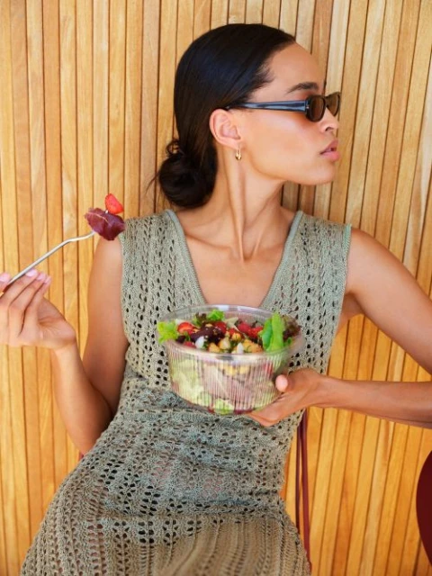 A young woman sits in front of a wooden wall, eating a salad. She's wearing sunglasses and a knit dress in a sunny, outdoor setting.