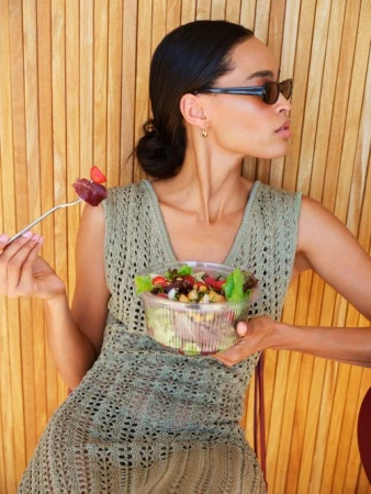 A young woman sits in front of a wooden wall, eating a salad. She's wearing sunglasses and a knit dress in a sunny, outdoor setting.
