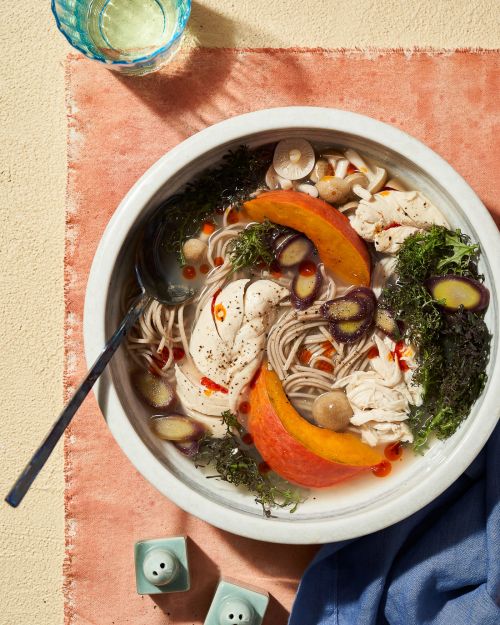 Overhead shot of a bowl of Asian-style soup with noodles, chicken, vegetables, and herbs on a peach-colored linen surface with salt shakers.