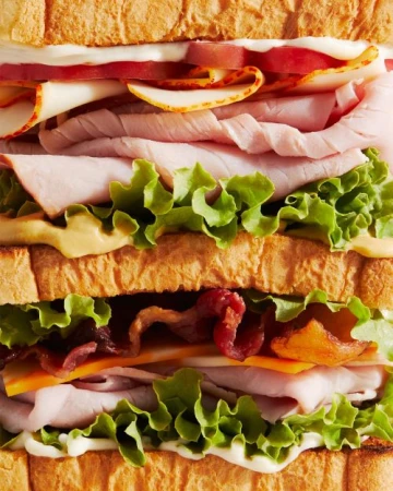 Overhead shot of a club sandwich. The sandwich is composed of toasted white bread, lettuce, tomato, bacon, cheese, ham, and turkey.