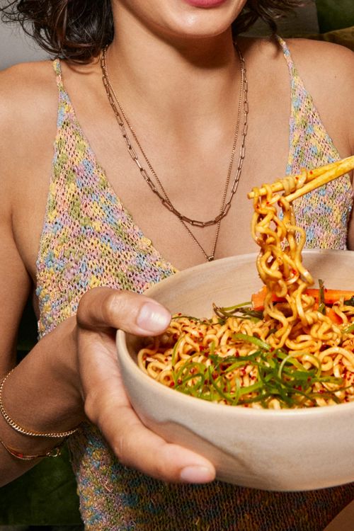 Close-up of woman holding a bowl of ramen with chopsticks, wearing a colorful top and gold necklaces. Food and fashion lifestyle.