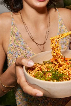 Close-up of woman holding a bowl of ramen with chopsticks, wearing a colorful top and gold necklaces. Food and fashion lifestyle.