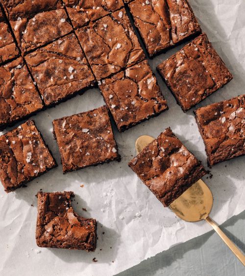 Overhead shot showcases a batch of freshly baked, sea-salt-sprinkled chocolate brownies served on parchment paper with a golden spatula.