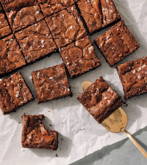 Overhead shot showcases a batch of freshly baked, sea-salt-sprinkled chocolate brownies served on parchment paper with a golden spatula.