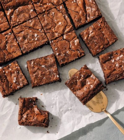Overhead shot showcases a batch of freshly baked, sea-salt-sprinkled chocolate brownies served on parchment paper with a golden spatula.