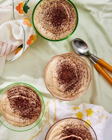 Overhead shot of four small bowls of mousse topped with cocoa powder, spoons, and a floral patterned cloth on a green surface.