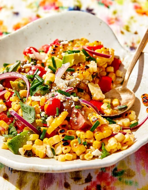 Overhead shot of corn salad in a white bowl, featuring grilled corn, tomatoes, avocado, red onion, herbs, and a golden spoon.