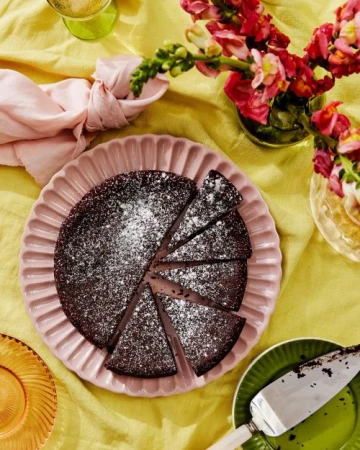 Overhead shot of a sliced chocolate cake dusted with powdered sugar on a pink scalloped plate surrounded by flowers and yellow tablecloth.