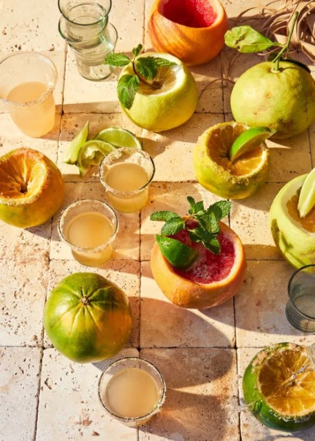 Overhead shot of citrus fruit drinks and citrus halves on a tiled surface, with mint leaves and salt-rimmed glasses.