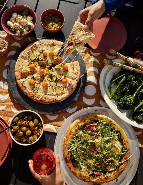 Overhead shot of two pizzas, one being served, surrounded by bowls of appetizers and drinks, creating a vibrant food spread for a meal.