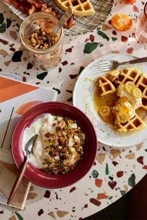 Overhead shot of breakfast food on a terrazzo table: waffles with citrus, yogurt and granola bowl, bacon, and a glass of beverage.