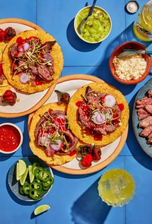 Overhead shot of multiple plates of tostadas topped with steak, onions, and sprouts with a side of limes, jalapenos, and sauces.