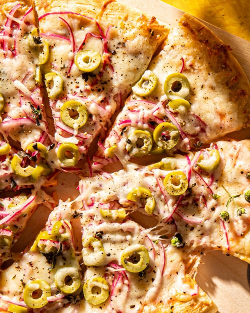 Overhead shot of a sliced pizza with melted cheese, red onions, and green olives, presented on a light surface with a yellow backdrop.