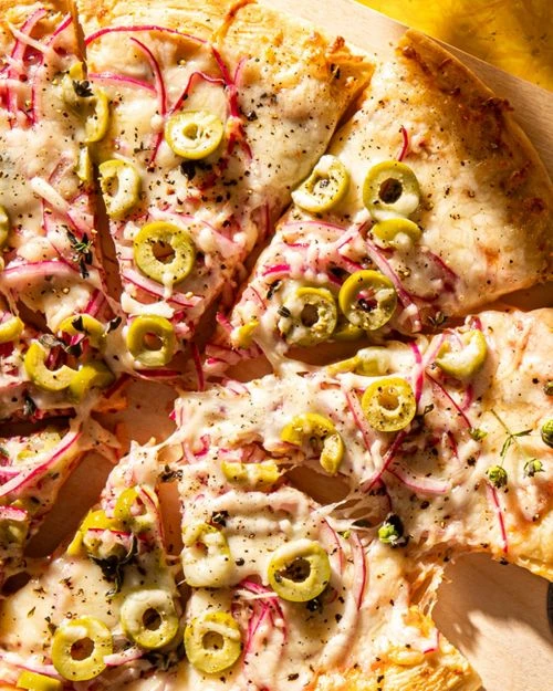 Overhead shot of a sliced pizza with melted cheese, red onions, and green olives, presented on a light surface with a yellow backdrop.
