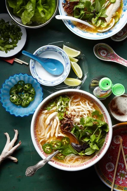 Overhead shot of bowls of pho with chicken, broth, noodles, garnishes, and spices on a dark green surface, next to spoons, chopsticks, and garnishes.