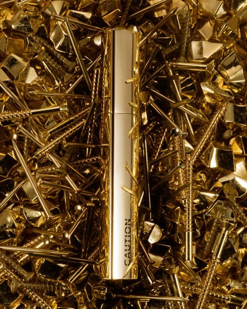 Still life shot of a tube of gold cosmetic product surrounded by a pile of golden metal hardware like nails and screws.