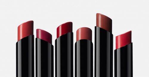 Several lipsticks are displayed against a light background. The lipsticks have black tubes and a range of red and brown shades.