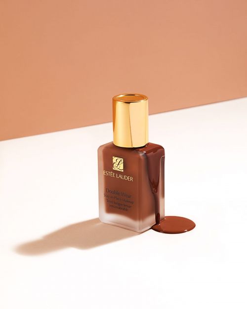 Estée Lauder Double Wear foundation in a glass bottle with a gold lid and brown liquid makeup against a two-toned background.
