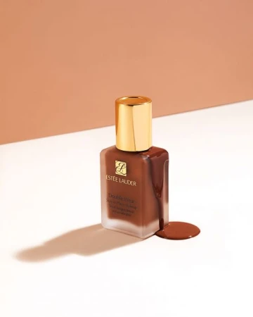 Estée Lauder Double Wear foundation in a glass bottle with a gold lid and brown liquid makeup against a two-toned background.