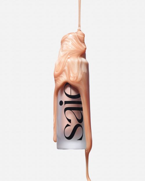 Cosmetic bottle with shiny, light-peach-colored liquid cascading down the side against a clean, bright, solid white backdrop.