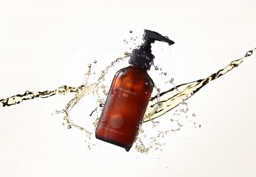 Product shot of an amber-colored bottle of skincare product with a black pump, surrounded by splashing clear and amber-colored liquid.
