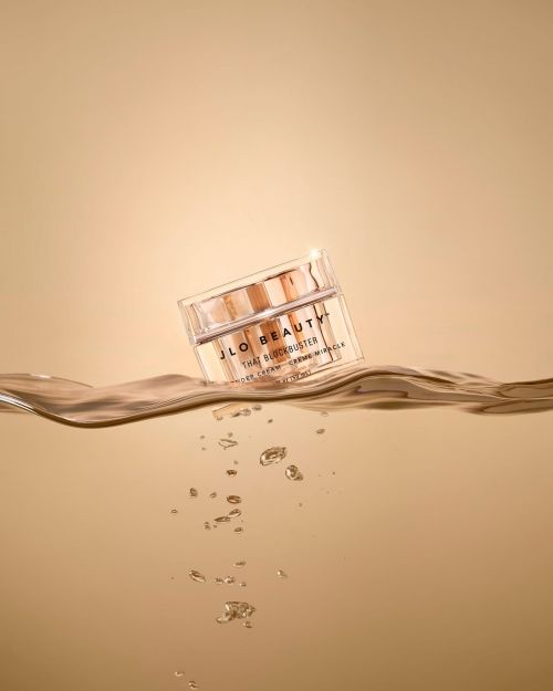 A jar of JLO Beauty cream on a surface of rippled water with bubbles, set against a soft beige background in a studio setting.
