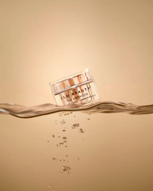 A jar of JLO Beauty cream on a surface of rippled water with bubbles, set against a soft beige background in a studio setting.