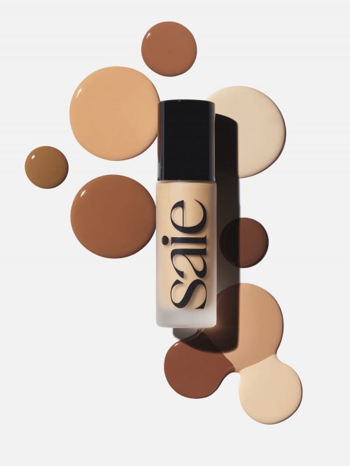 Studio shot of a bottle of Saie foundation surrounded by drops of foundation in varying skin tone shades against a white background.
