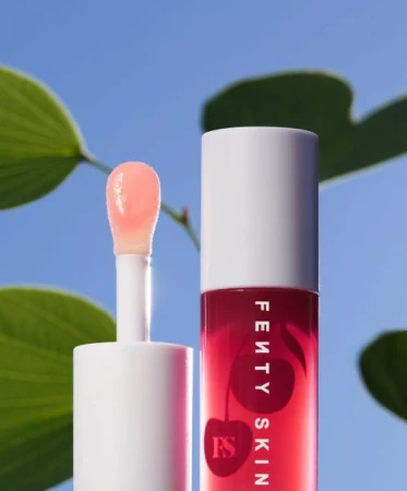 Fenty Skin Cherry Treat lip conditioning butter is positioned against a bright blue sky and out-of-focus green foliage backdrop.