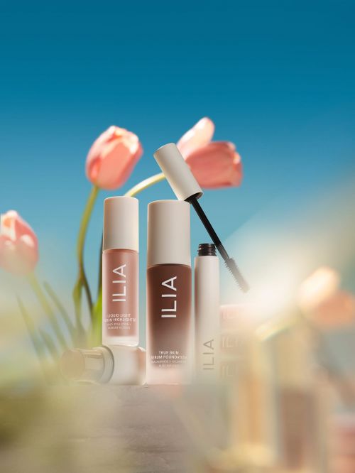 Cosmetic products from Ilia including foundation and serum are arranged against a bright blue sky with blurred pink tulips.