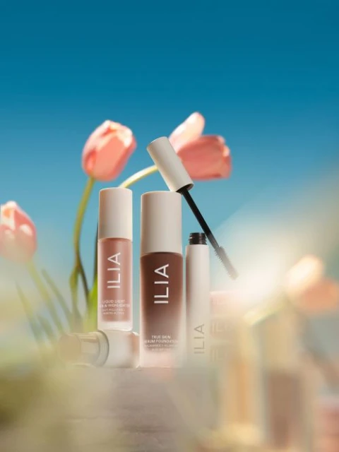 Cosmetic products from Ilia including foundation and serum are arranged against a bright blue sky with blurred pink tulips.