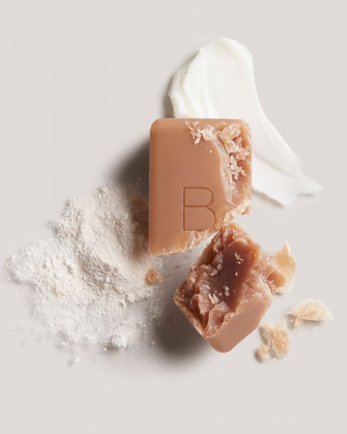 Close-up of tan-colored soap bar with carved letter 'B' with smear of white cream and white powder against a neutral background.