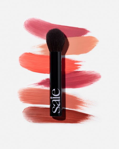 Makeup brush with black handle on white background next to blush swatches in red and peach tones. Beauty product and cosmetics.
