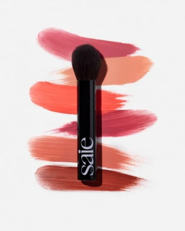 Makeup brush with black handle on white background next to blush swatches in red and peach tones. Beauty product and cosmetics.