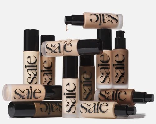 Stacked Saie brand foundation bottles in various shades with minimalist labels and black pump caps against a neutral backdrop.