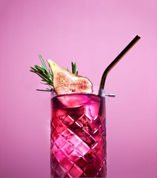 Refreshing pink cocktail in a textured glass with fig and rosemary garnish, complete with a metal straw against a pink background.