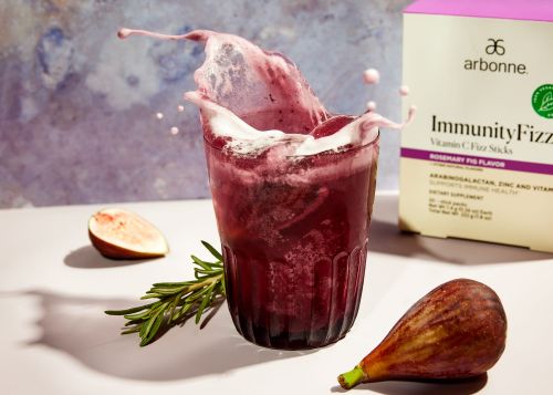 A vibrant purple beverage splashes from a glass, next to figs, rosemary sprigs, and an Arbonne Immunity Fizz box on a white surface.