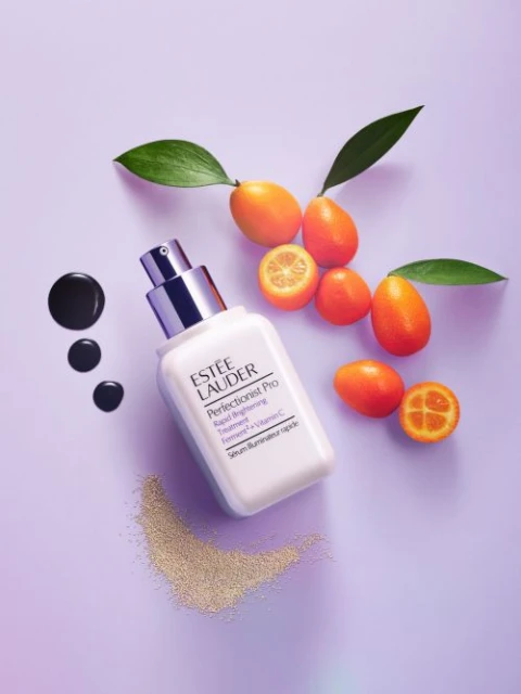 Estee Lauder serum bottle sits on a lavender surface with kumquats, green leaves, and cosmetic accents, promoting beauty and skincare.