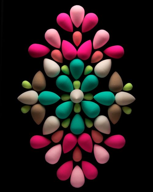 Eye-level, close-up studio shot of teardrop makeup sponges in pink, white, tan, green, and turquoise arranged in symmetrical floral pattern on black.
