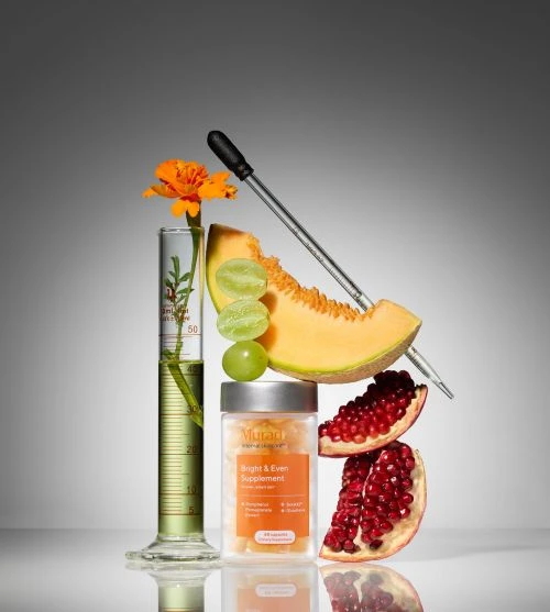 Still life composition of fruits, flower, supplement bottle, and lab equipment in a studio setting with neutral gray background.