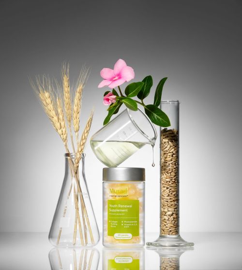 Still life shot of a bottle of youth renewal supplement, flowers, wheat stalks, and lab beakers on a white surface with a gray background.