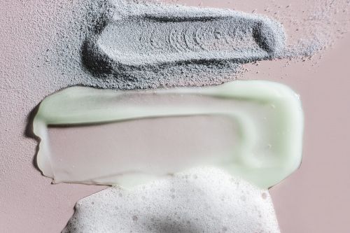 Close-up of cosmetic textures in grey powder, pale green gel, and white foam arranged on a pink background in a studio setting.