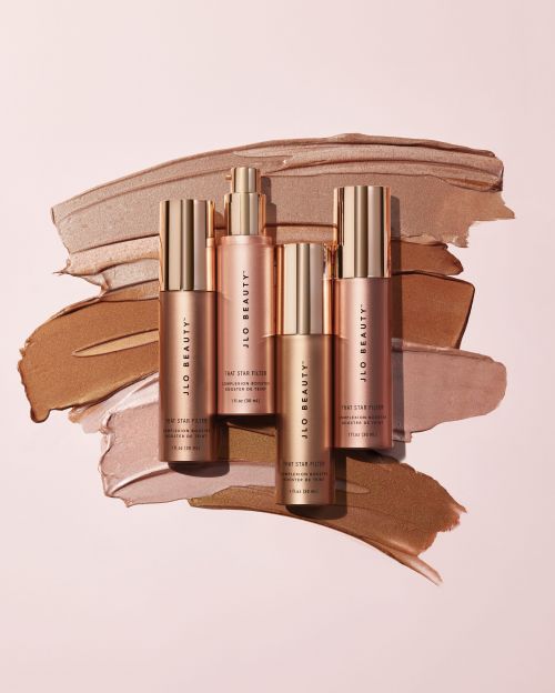 Four JLo Beauty serum bottles arranged on a soft pink surface with beige and brown cosmetic swatches for a beauty product shot.